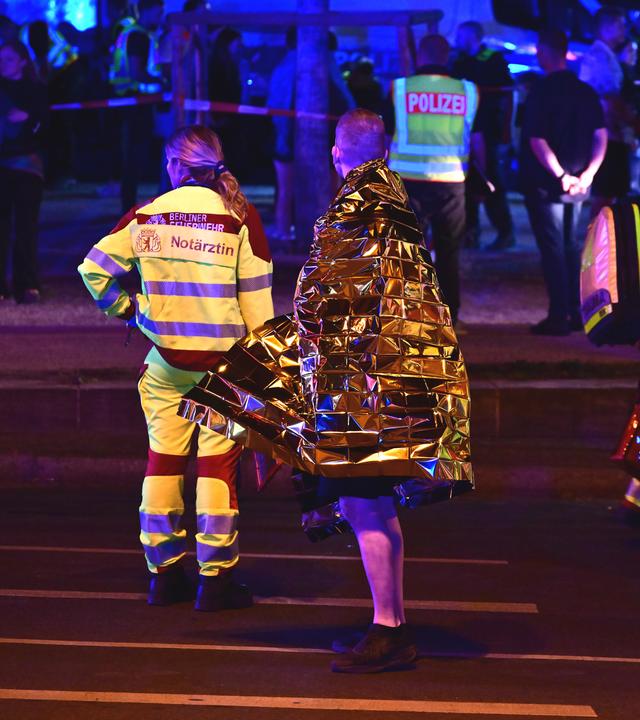 Eine in eine Rettungsdecke gehüllte Person steht neben Einsatzkräften der Feuerwehr nach dem Abbruch des Christopher Street Day (CSD)