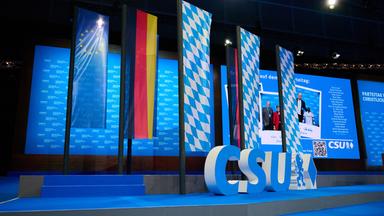 Ein großes CSU-Logo steht auf der Bühne, im Hintergrund drei blau-weiß-karierte Bayern-Fahnen, eine Deutschlandflagge und eine EU-Flagge.