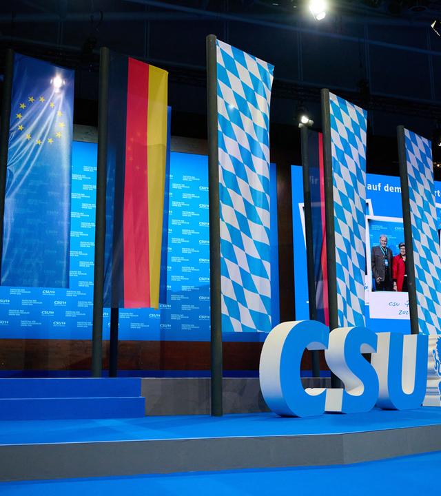 Ein großes CSU-Logo steht auf der Bühne, im Hintergrund drei blau-weiß-karierte Bayern-Fahnen, eine Deutschlandflagge und eine EU-Flagge.