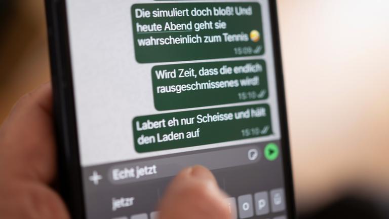 Eine Person hält ein Smartphone in der Hand, auf dessen Bildschirm beleidigende Nachrichten in einem Chat zu sehen sind (gestellte Szene).