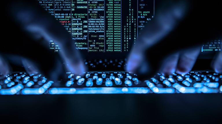 Symbolbild: Ein Mann tippt auf der beleuchteten Tastatur eines Laptops. Auf dem Bildschirm ist ein Hackerprogramm geöffnet. 