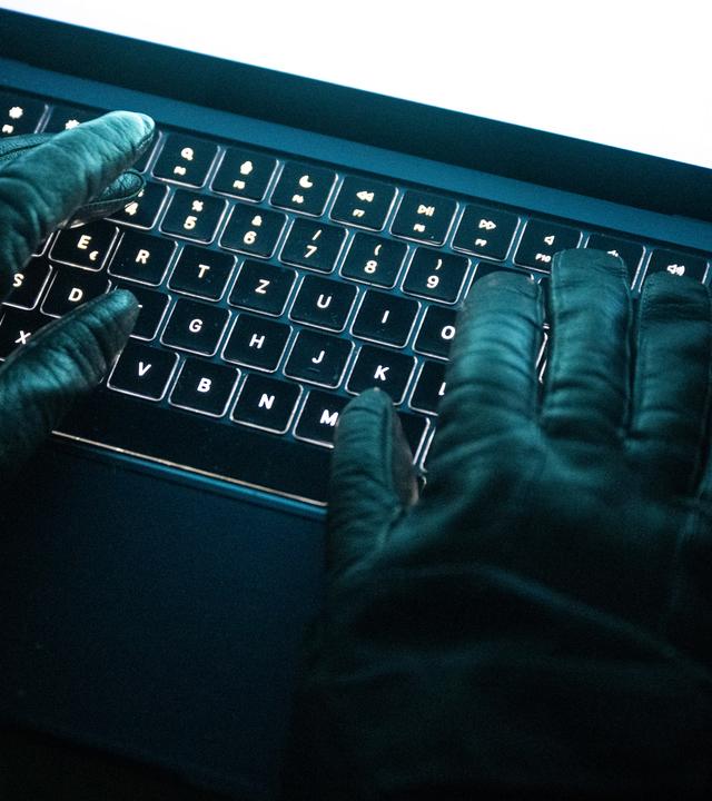 Eine Person tippt mit behandschuhten Händen auf einer beleuchteten Laptop-Tastatur. (Gestellte Szene)