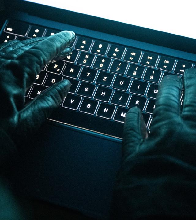 Eine Person tippt mit behandschuhten Händen auf einer beleuchteten Laptop-Tastatur. 