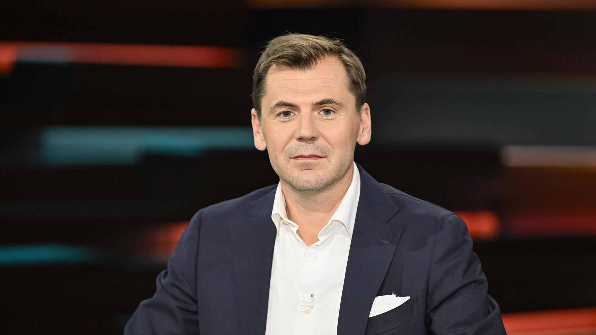 Nahost-Experte Daniel Gerlach in einem dunkelblauen Anzug mit weißem Hemd vor einem verschwommenen Hintergrund mit orange-dunkelblauem Licht bei Markus Lanz.