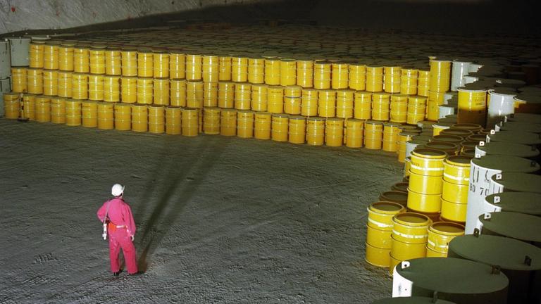  Blick von oben auf große, gelbe Fässer, die in drei Reihen übereinandergestapelt sind; sie stehen in einer unterirdischen Halle und füllen den gesamten hinteren Teil des Raumes. Davor steht ein Mann in roter Arbeitsmontur und Helm mit dem Rücken zur Kamera.