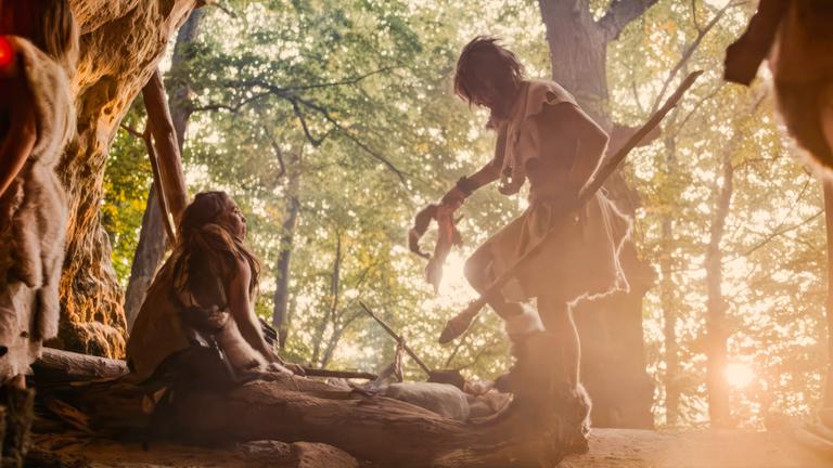 Spielszene: In einer Höhle in einem Wald halten sich mehrere Steinzeitmenschen auf. Im Hintergrund stehen viele Bäume durch die die Sonne scheint.