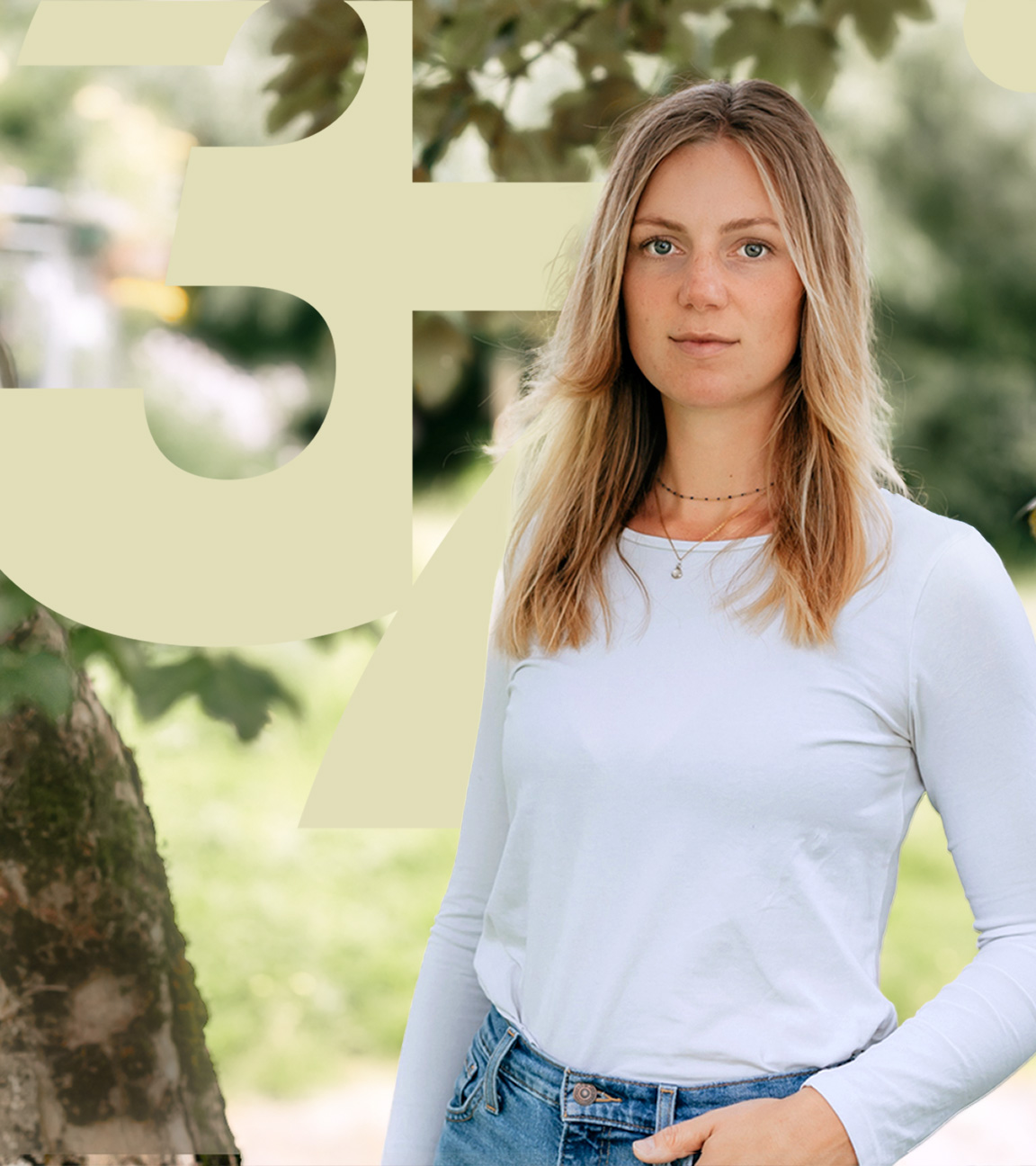 Mara steht in der Natur vor einem Baum und schaut in die Kamera. Eine Hand hat sie leicht in ihre Hosentasche gesteckt. 