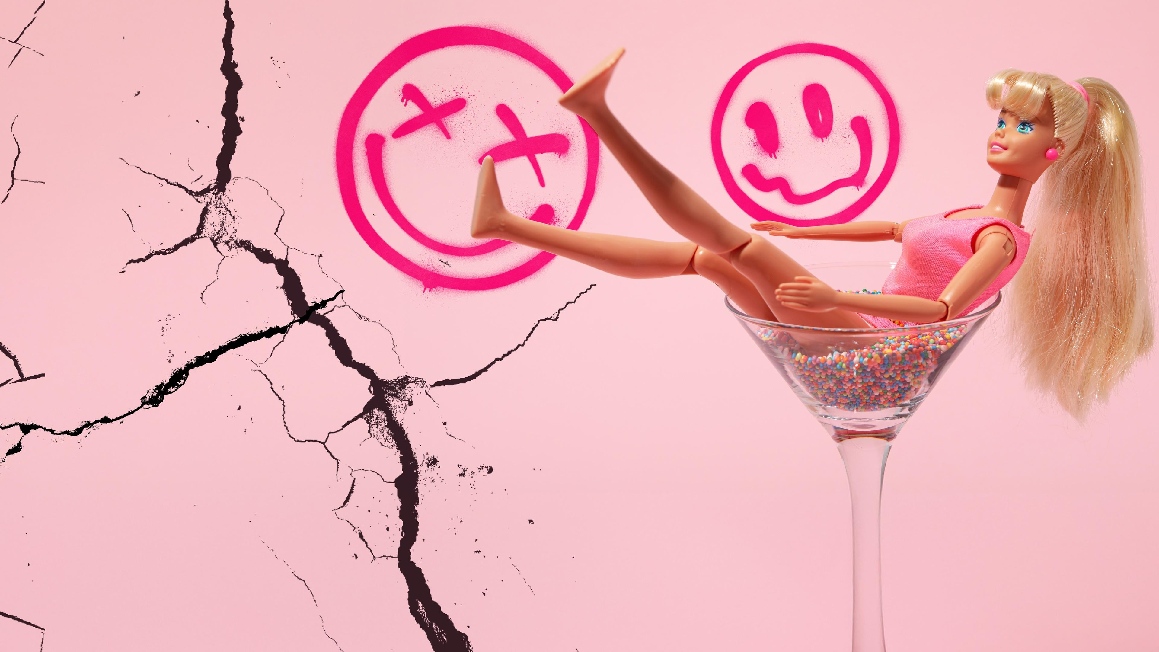 Eine Barbiepuppe liegt in einem Cocktailglas. Der rosane Hintergrund hat Risse und aufgesprühte Graffiti-Smileys
