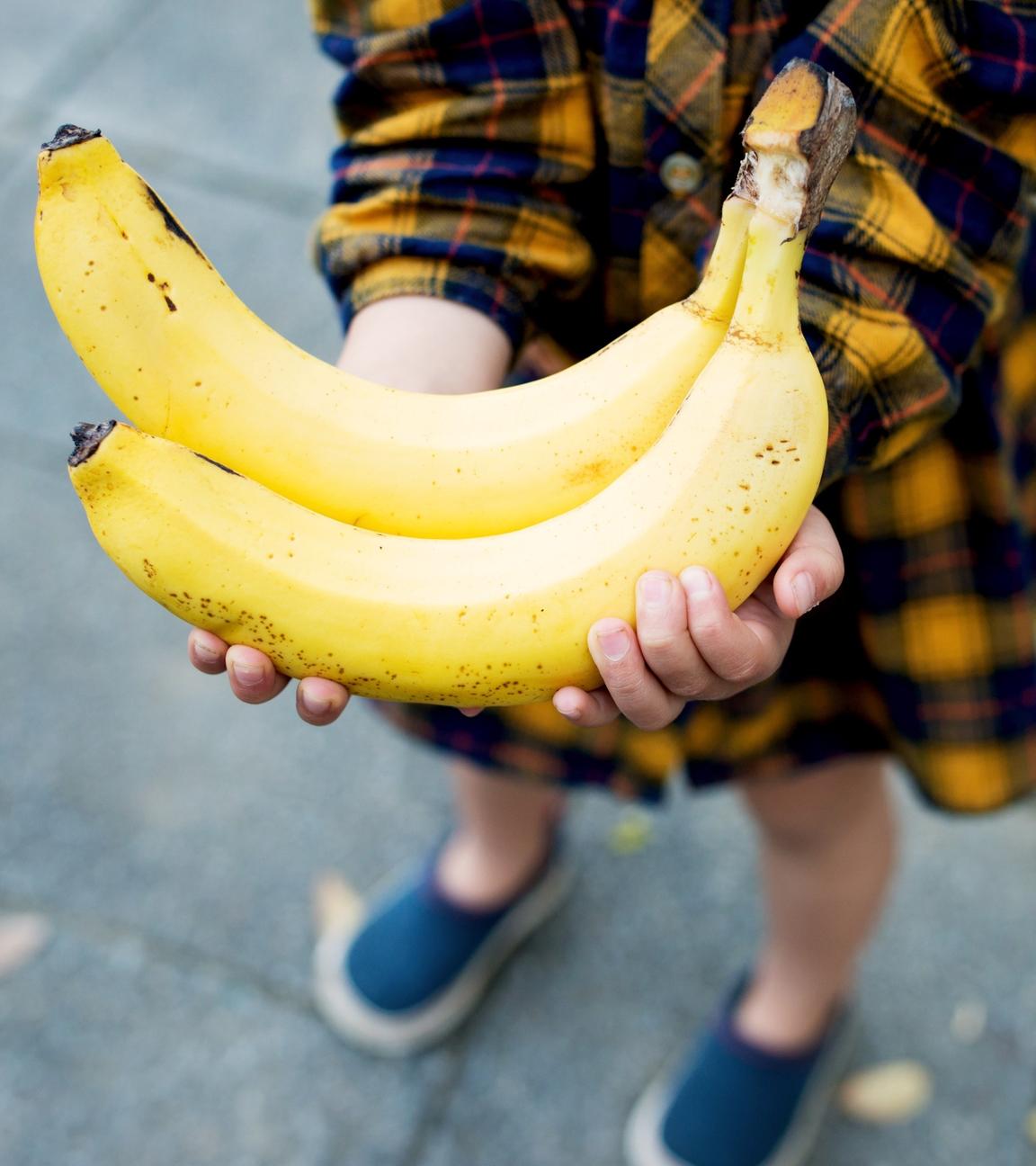 Blick von schräg oben auf zwei gelbe Bananen in den Händen eines Kindes.