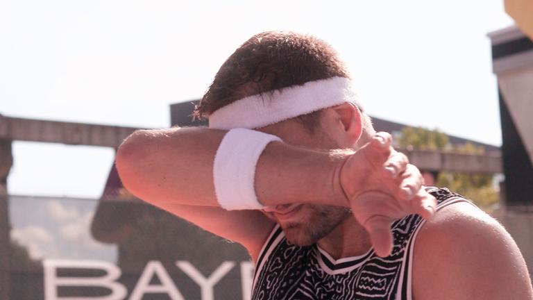 Mann im Sportoutfit mit Schweißbändern an Kopf und Handgelenk. Er wischt sich mit einem Schweißband durch das Gesicht.