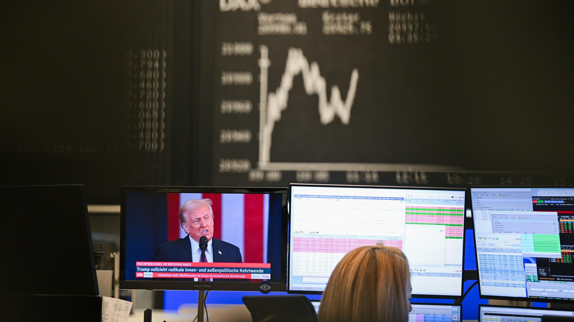 Die Dax-Kurve zeigt im Handelssaal der Frankfurter Wertpapierbörse Auf- und Abwärtsbewegungen, während auf einem Bildschirm der neue US-Präsident Donald Trump zu sehen ist.