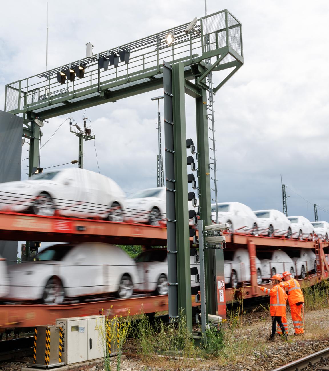 Foto eines vorbeifahrenden, unscharfen Güterzugs der DB Cargo der mit weiß folierten Autos beladen ist, zwei Mitarbeiter in Sicherheitswesten stehen am Gleis.