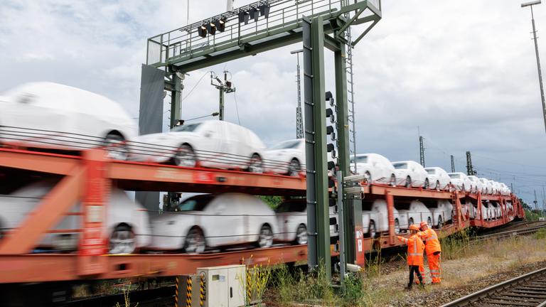 Foto eines vorbeifahrenden, unscharfen Güterzugs der DB Cargo der mit weiß folierten Autos beladen ist, zwei Mitarbeiter in Sicherheitswesten stehen am Gleis.