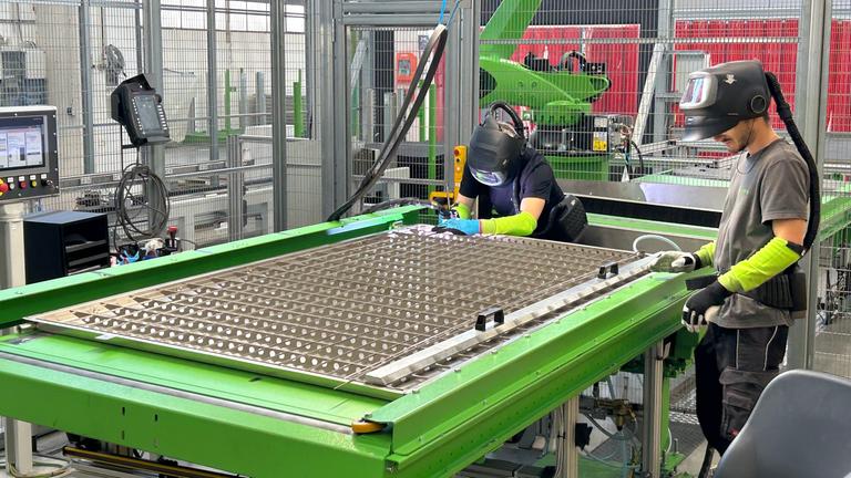 Mitarbeiter mit Schutzhelmen arbeiten an einem Tisch: Bei De Nora werden Elektroden behandelt, die für die Wasserstoffgewinnung nötig sind.