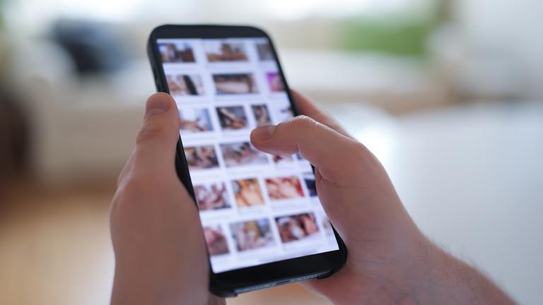 Ein junger Mann schaut sich auf einem Smartphone eine Porno-Internetseite an (gestellte Szene).