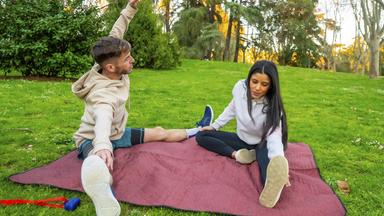Junges Paar in Sportkleidung dehnt sich gemeinsam auf einer kastanienbraunen Picknickdecke in einem sonnigen Park