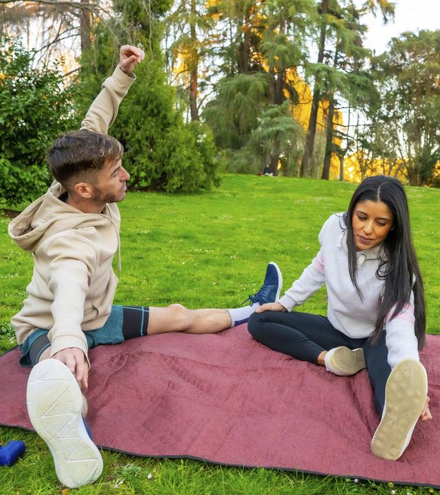 Junges Paar in Sportkleidung dehnt sich gemeinsam auf einer kastanienbraunen Picknickdecke in einem sonnigen Park