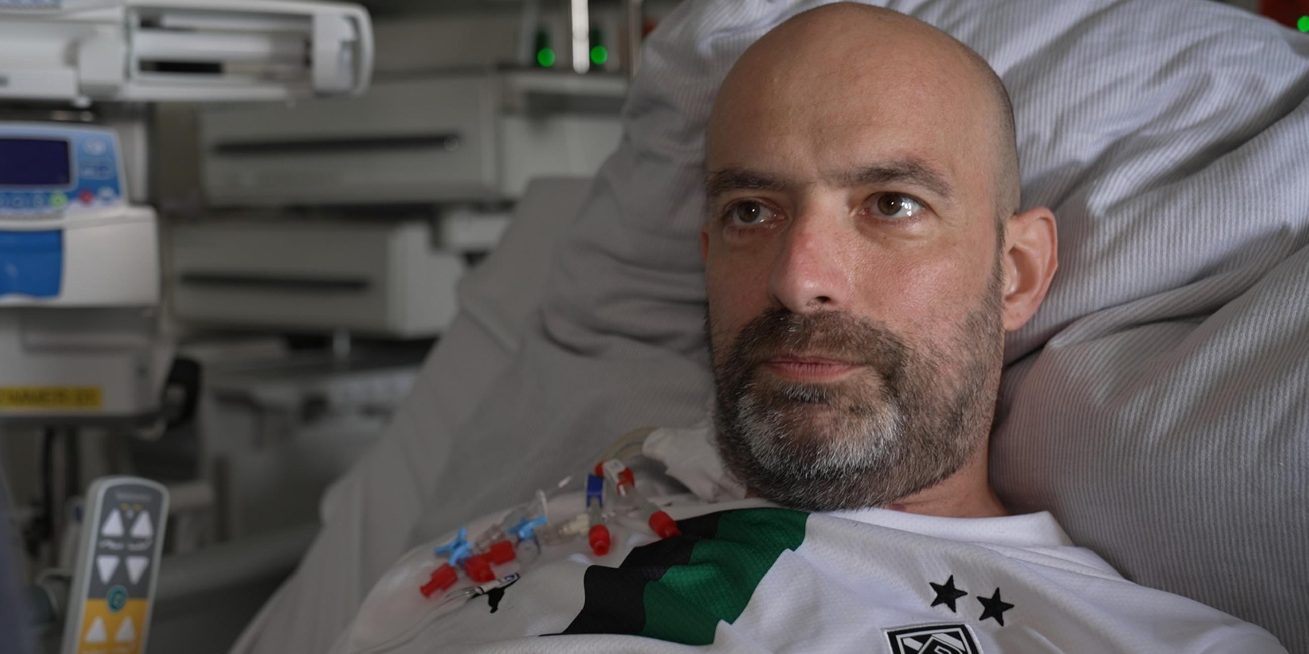 Dirk ist mittig im Bild im Porträt zu sehen. Er sitzt im Krankenhausbett, hinter ihm sind medizinische Geräte zu erkennen. Er schaut rechts an der Kamera vorbei.