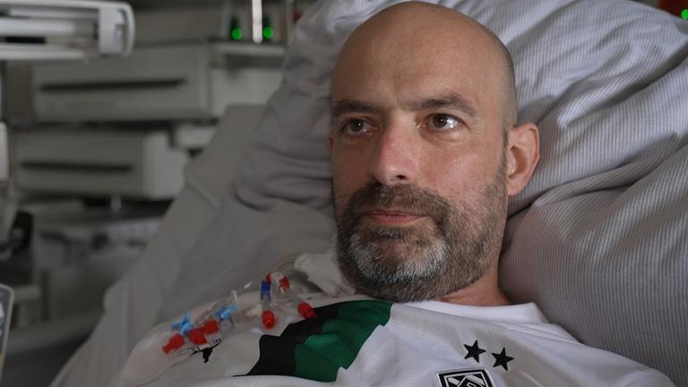 Dirk ist mittig im Bild im Porträt zu sehen. Er sitzt im Krankenhausbett, hinter ihm sind medizinische Geräte zu erkennen. Er schaut rechts an der Kamera vorbei.