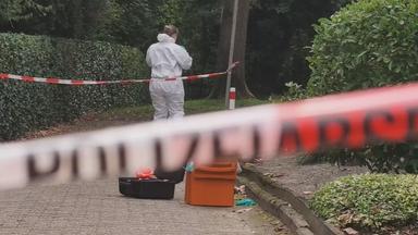 Auf dem Bild ist zu sehen: Ein rot-weißes Absperrband verläuft unscharf quer durch den Vordergrund. Dahinter steht eine Person in einem weißen Schutzanzug mit dem Rücken zur Kamera