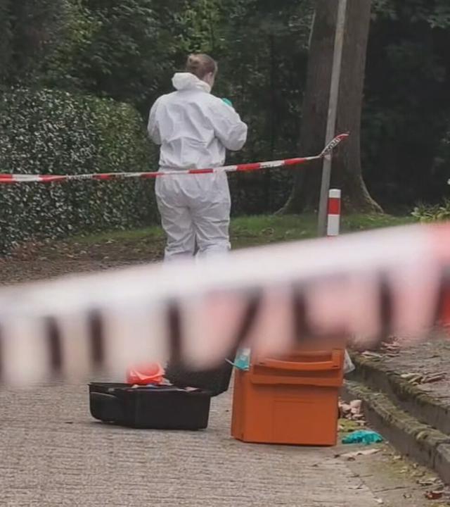 Auf dem Bild ist zu sehen: Ein rot-weißes Absperrband verläuft unscharf quer durch den Vordergrund. Dahinter steht eine Person in einem weißen Schutzanzug mit dem Rücken zur Kamera