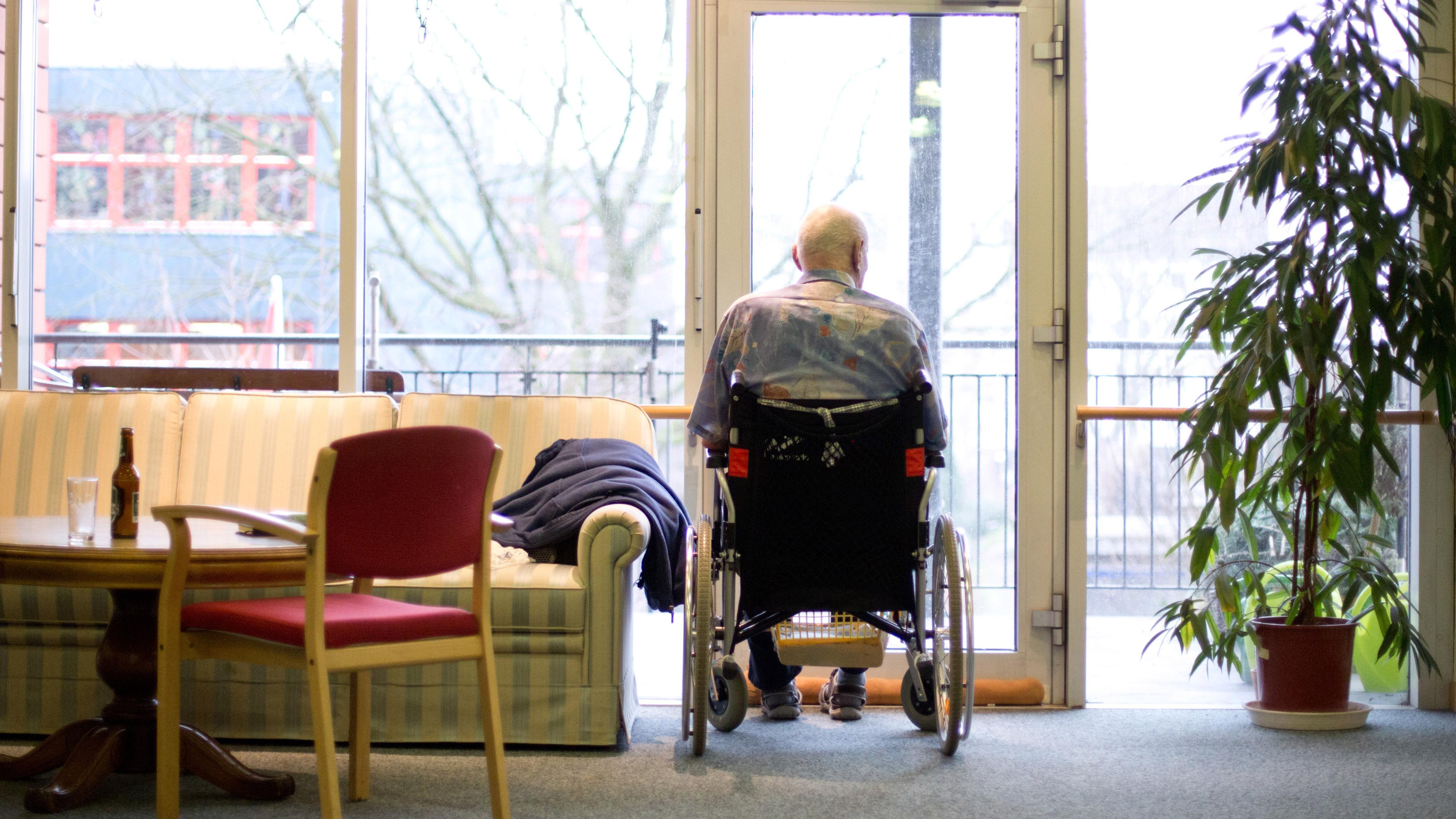 Ein alter Mann sitzt im Rollstuhl am Fenster und blickt heraus.