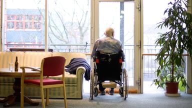 Ein alter Mann sitzt im Rollstuhl am Fenster und blickt heraus.