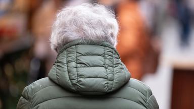 Eine ältere Frau trägt eine grüne Winterjacke. Man sieht sie nur von hinten
