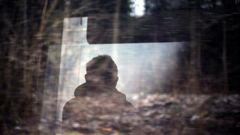 Ein Mann blickt aus dem Fenster in einen Wald. Die Bäume haben keine Blätter mehr und alles ist sehr trist.