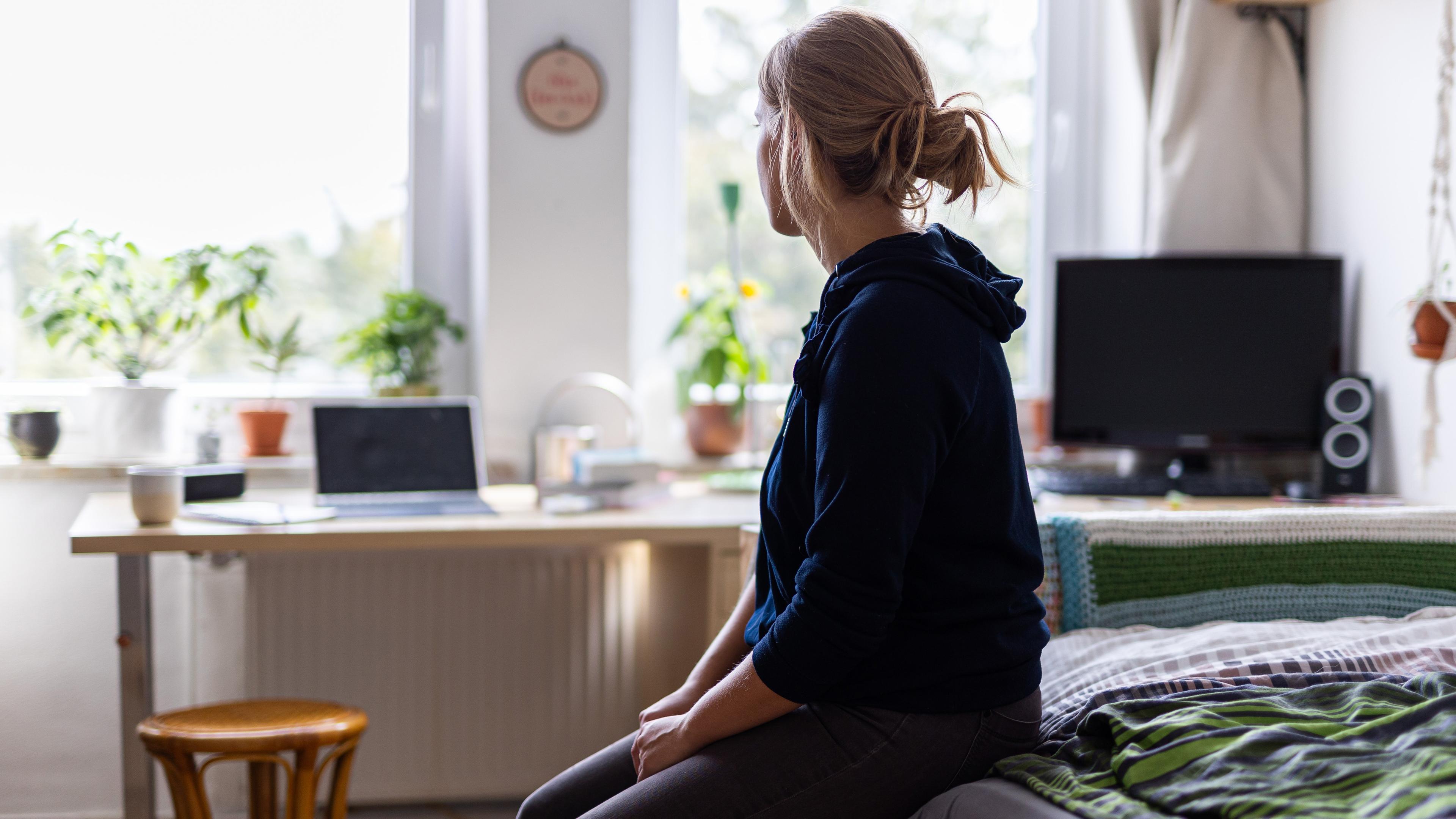 ILLUSTRATION - Eine Studentin sitzt auf dem Bett in ihrem WG-Zimmer und schaut aus dem Fenster (gestellte Szene)