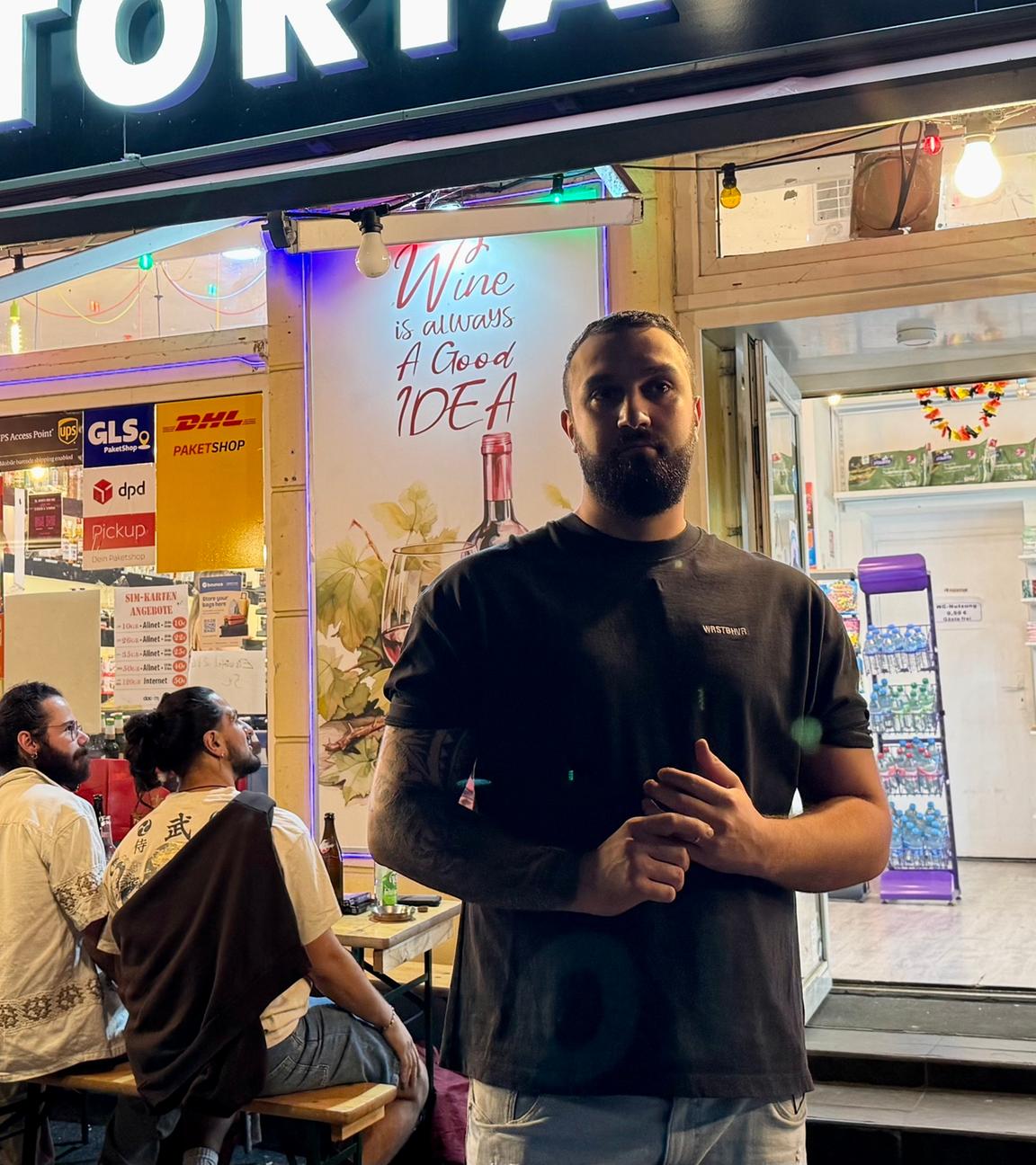 Serdar steht spät in der Nacht vor seinem Kiosk. Im Hintergrund sitzen zahlreiche Gäste auf Bänken, vertieft in Gespräche.