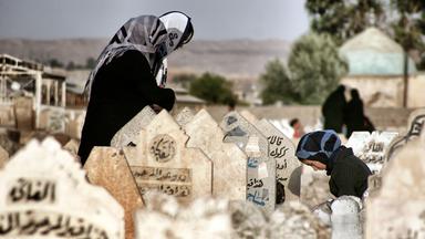 "Der Preis des Krieges: Irak": Drei Frauen mit Kopftüchern stehen mit geneigten Köpfen um ein Grab auf einem Friedhof. Eine Frau kniet. Es sind arabische Inschriften auf den Grabsteinen zu lesen.