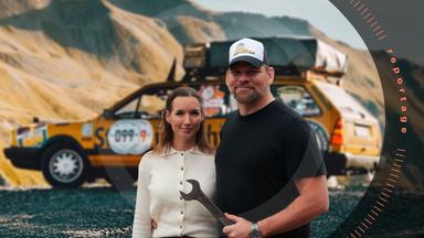 Hardy Leben und Freundin Jessica Hetke umarmen sich. Beide stehen vor ihrem zu einem Pannenhelfer-Auto umgebauten VW-Passat-Oldtimer. Hardy Leben hält einen großen Schraubenschlüssel in Hand. 