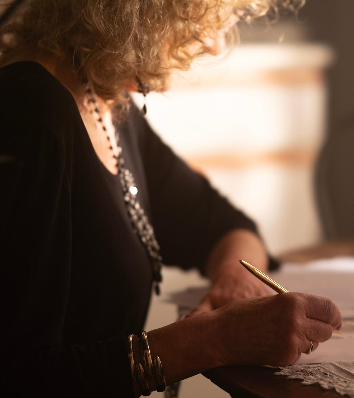 Eine Frau sitzt an einem Tisch vor einem Blatt Papier. In der rechten Hand hält sie einen Stift. Vor ihr steht ein gefülltes Weinglas
