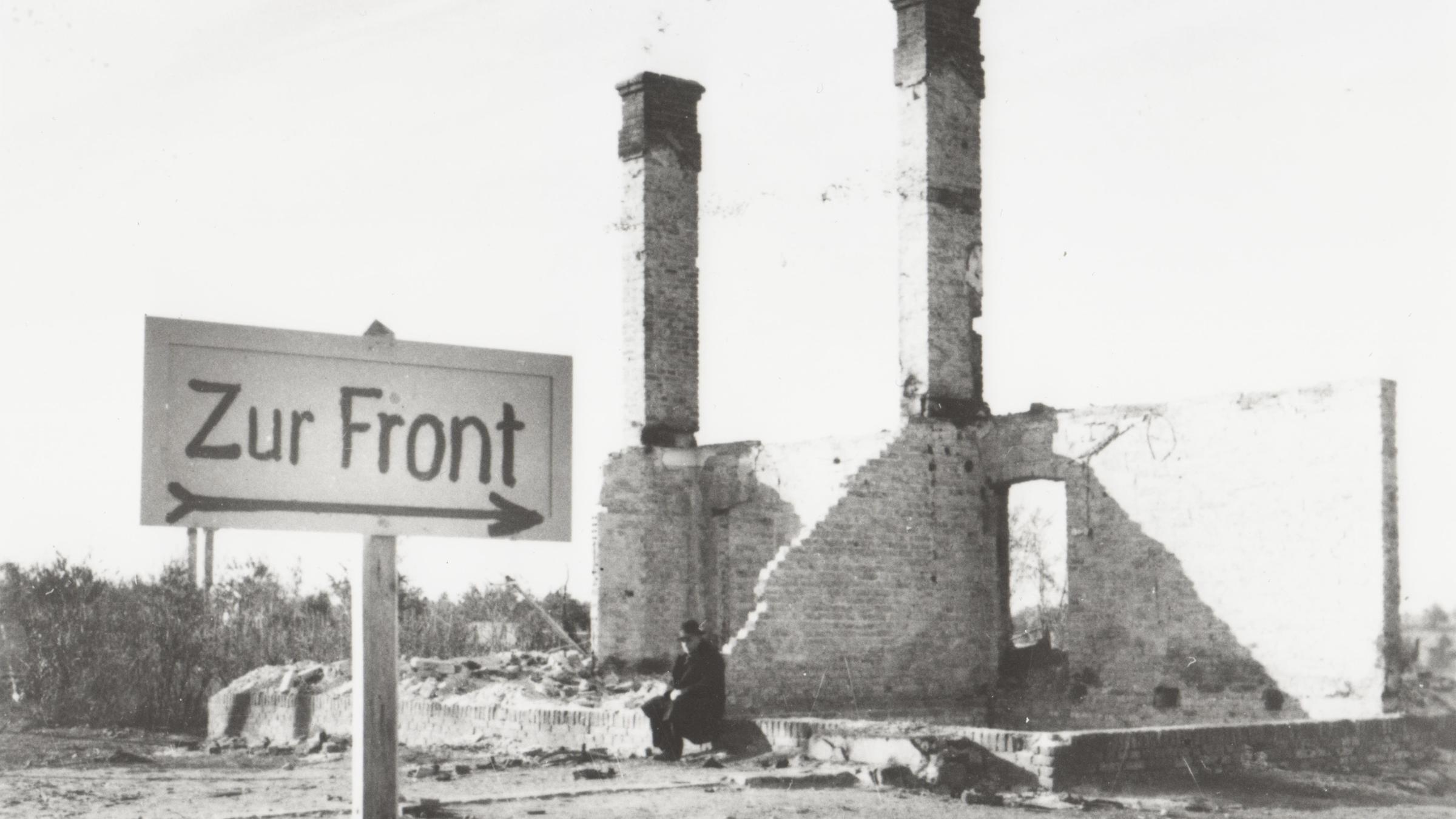 "Der Zweite Weltkrieg: Der Überfall": Ein Mann sitzt in den Ruinen eines durch den Krieg zerstörten Hauses in einer polnischen Ortschaft. Im Vordergrund ist ein Wegweiser mit der Aufschrift "Zur Front" errichtet worden.