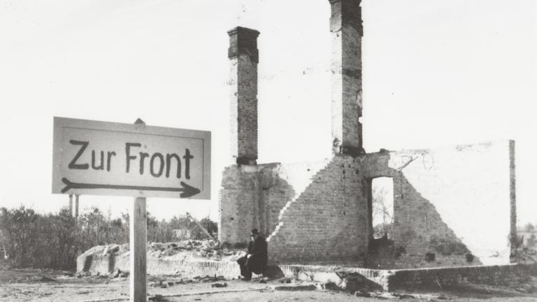 "Der Zweite Weltkrieg: Der Überfall": Ein Mann sitzt in den Ruinen eines durch den Krieg zerstörten Hauses in einer polnischen Ortschaft. Im Vordergrund ist ein Wegweiser mit der Aufschrift "Zur Front" errichtet worden.