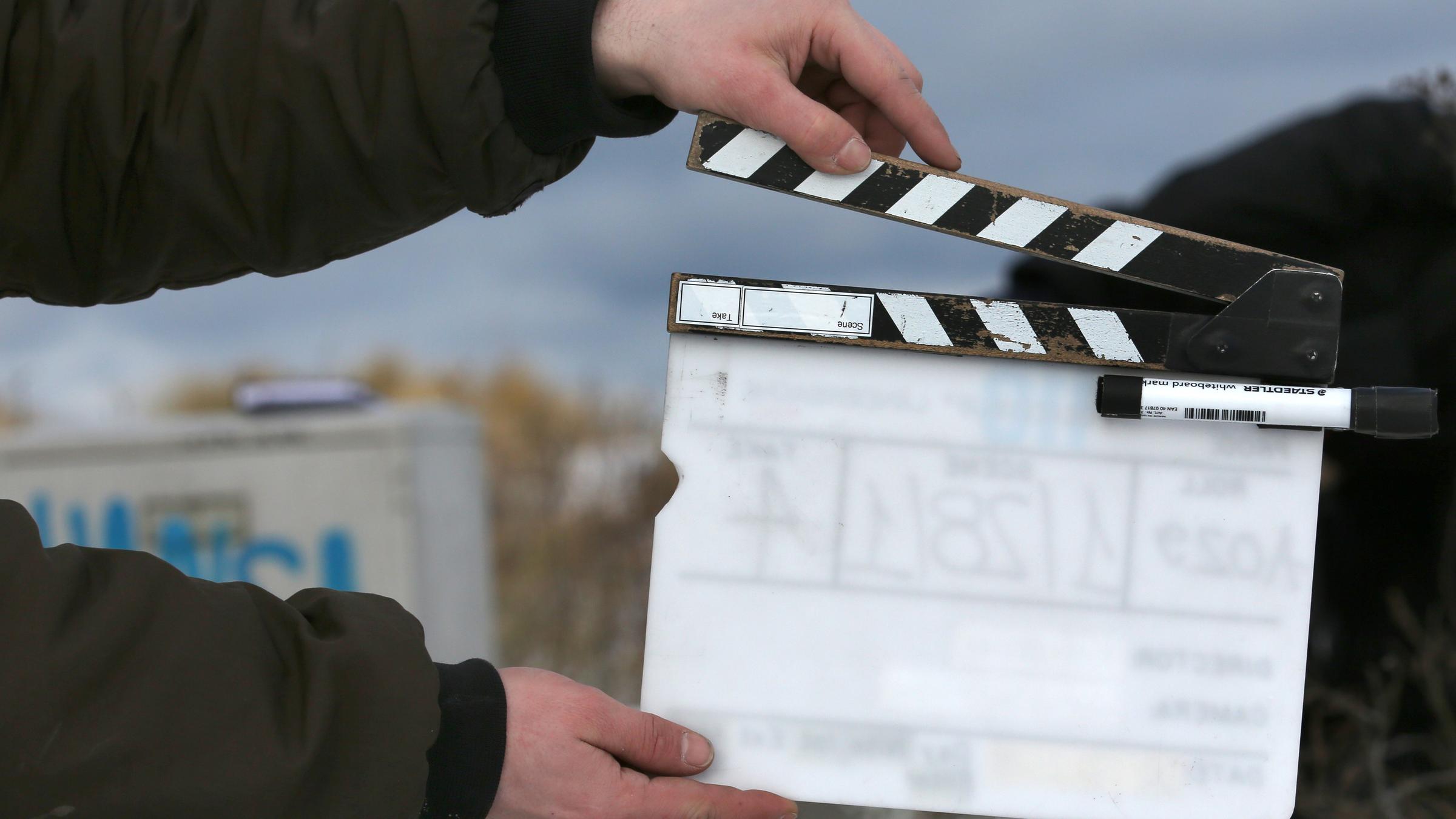 Eine Filmklappe wird von jemandem, dessen Arme nur zu sehen sind, vor das Set einer Produktion gehalten.