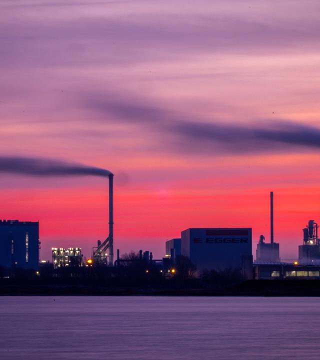 ARCHIV - 18.12.2025, Mecklenburg-Vorpommern, Wismar: Dampf steigt aus den Schornsteinen der Holzwerke am Seehafen, während die aufgehende Sonne den Himmel orangerot einfärbt.