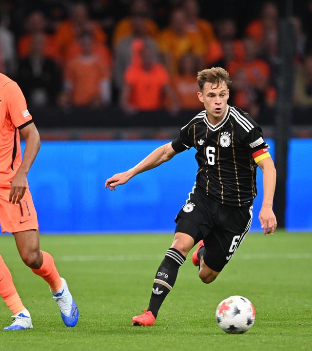 zwei Fußballer jagen dem Ball hinterher. Rechts steht Kimmich im schwarzen Trikot, links Cody Gakpo im orangen Trikot