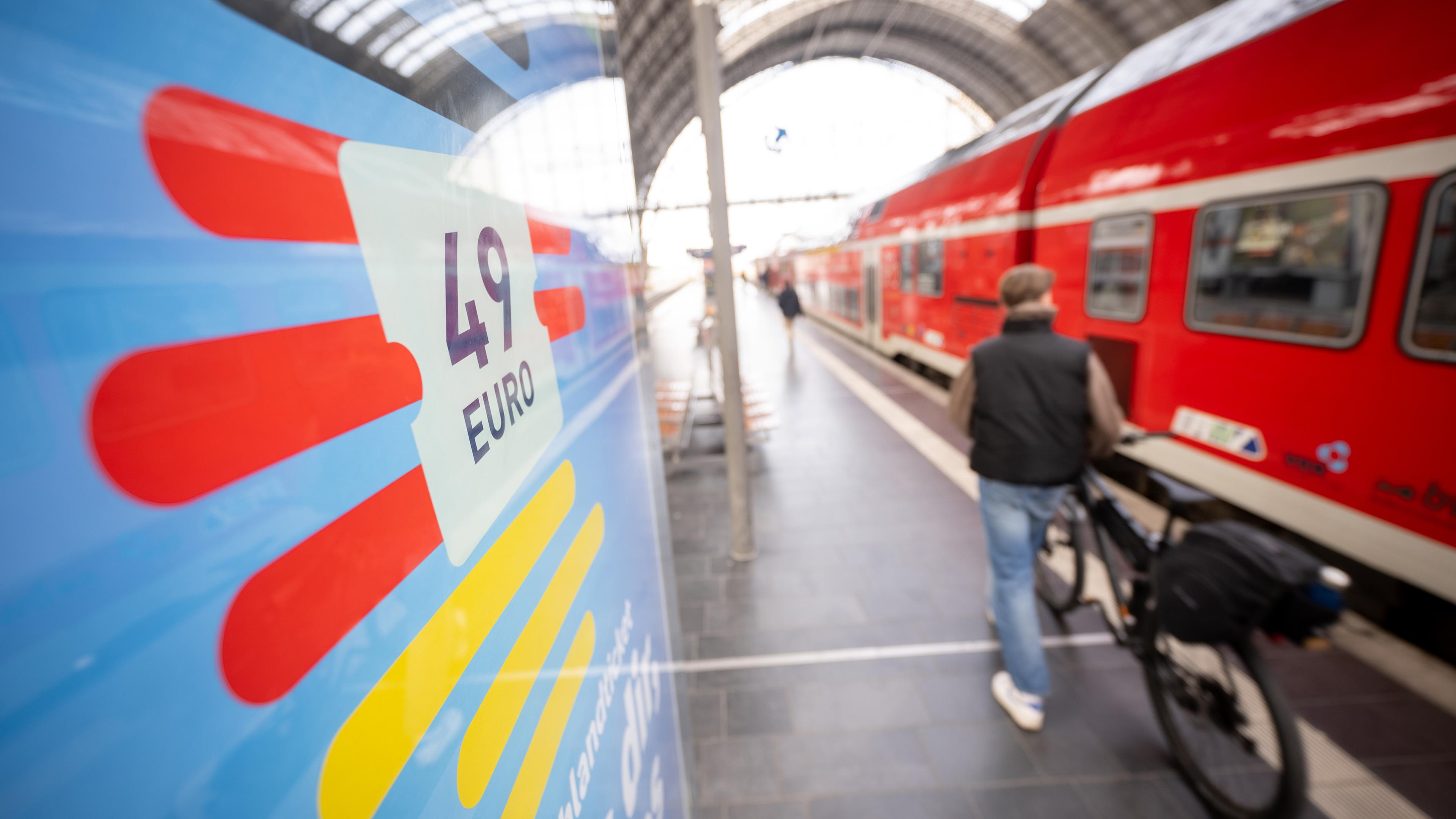 Ein Mann geht am Hauptbahnhof mit seinem Fahrrad zwischen der Werbung für das Deutschlandticket und einer Regionalbahn vorbei.