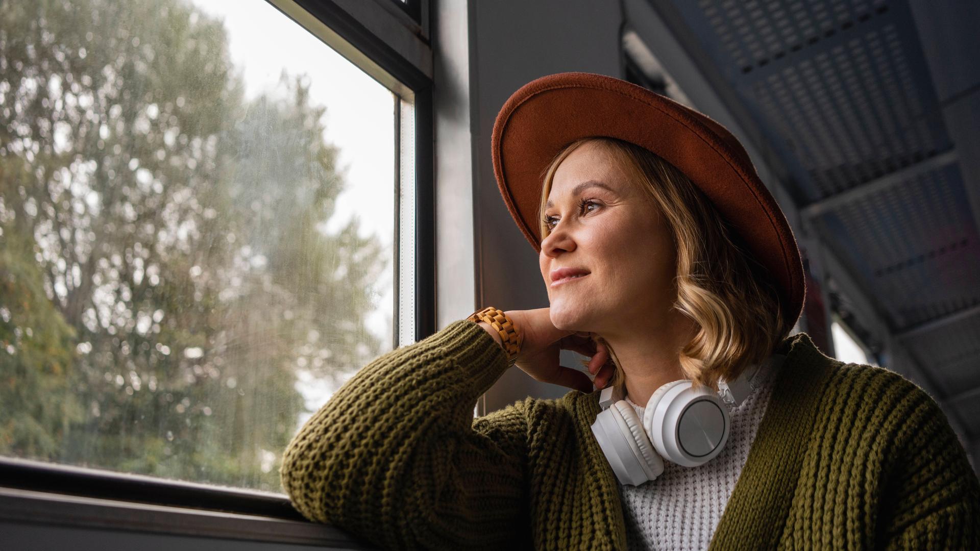 Eine Frau mit Hut und Kopfhörern, die sie um den Hals trägt, sitzt im Zug und blickt aus dem Fenster.