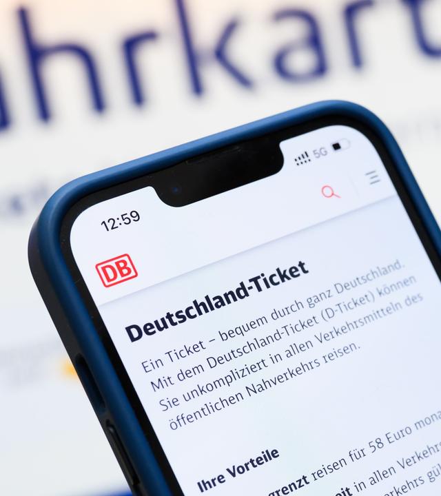 Eine Person hält am Hauptbahnhof ein Smartphone in der Hand, auf dessen Display eine Information zum Kauf des Deutschlandtickets zu sehen ist, während sie vor einem Fahrkartenautomaten steht. 