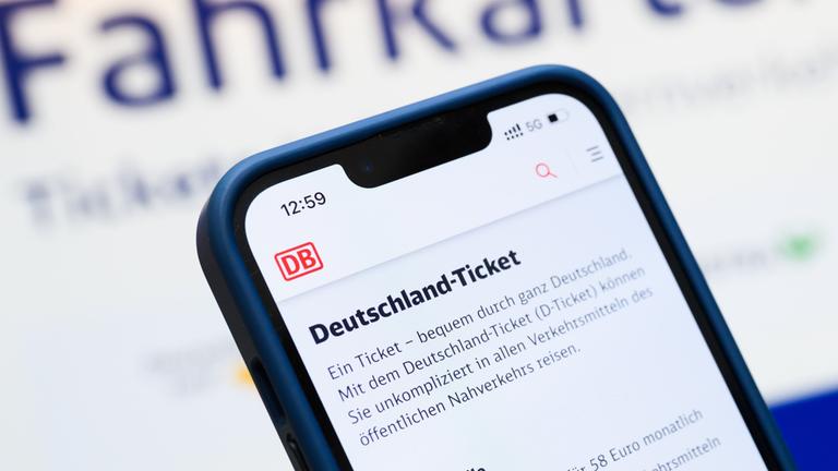 Eine Person hält am Hauptbahnhof ein Smartphone in der Hand, auf dessen Display eine Information zum Kauf des Deutschlandtickets zu sehen ist, während sie vor einem Fahrkartenautomaten steht. 