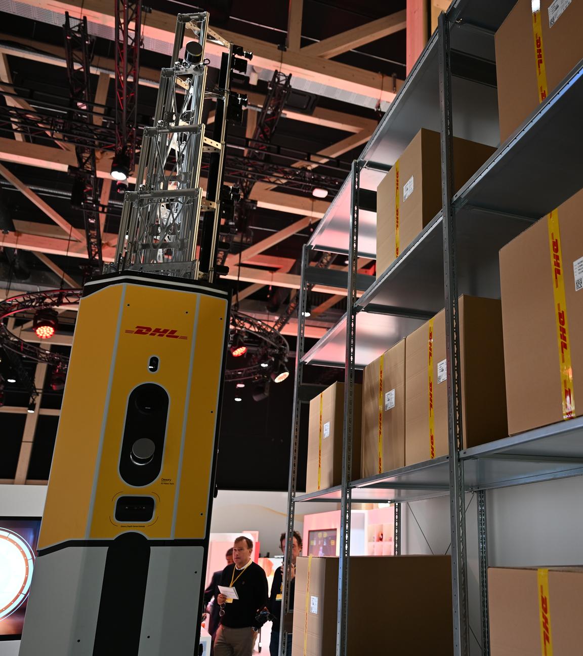 Ein Verteilerzentrum von DHL ist zu sehen. Es befinden sich einige Pakete im Zentrum und links von einem Regal voller Paketen befindet sich ein Roboter mit der Aufschrift "DHL". Dieser greift gerade zu einem Paket.
