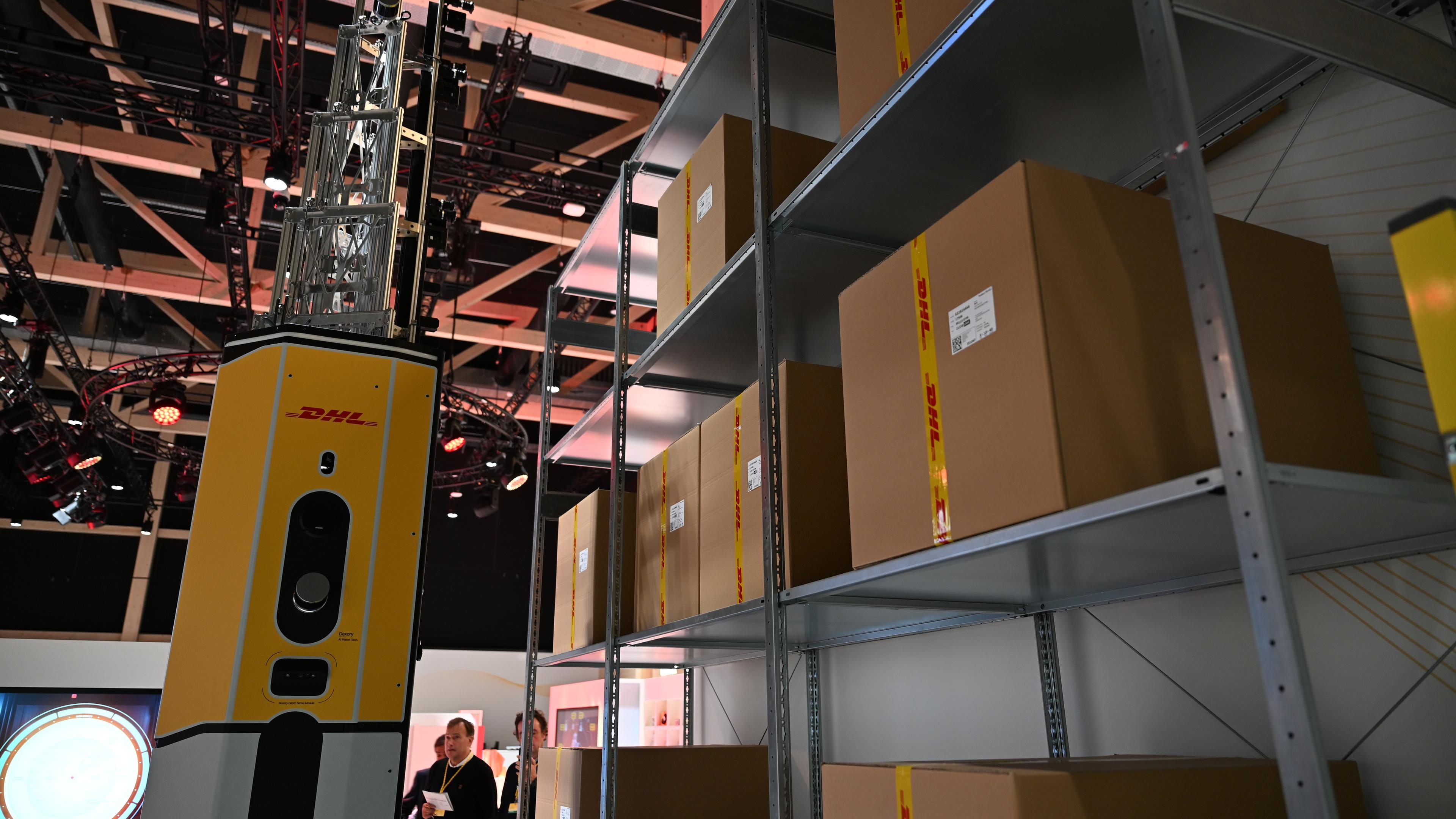 Ein Verteilerzentrum von DHL ist zu sehen. Es befinden sich einige Pakete im Zentrum und links von einem Regal voller Paketen befindet sich ein Roboter mit der Aufschrift "DHL". Dieser greift gerade zu einem Paket.