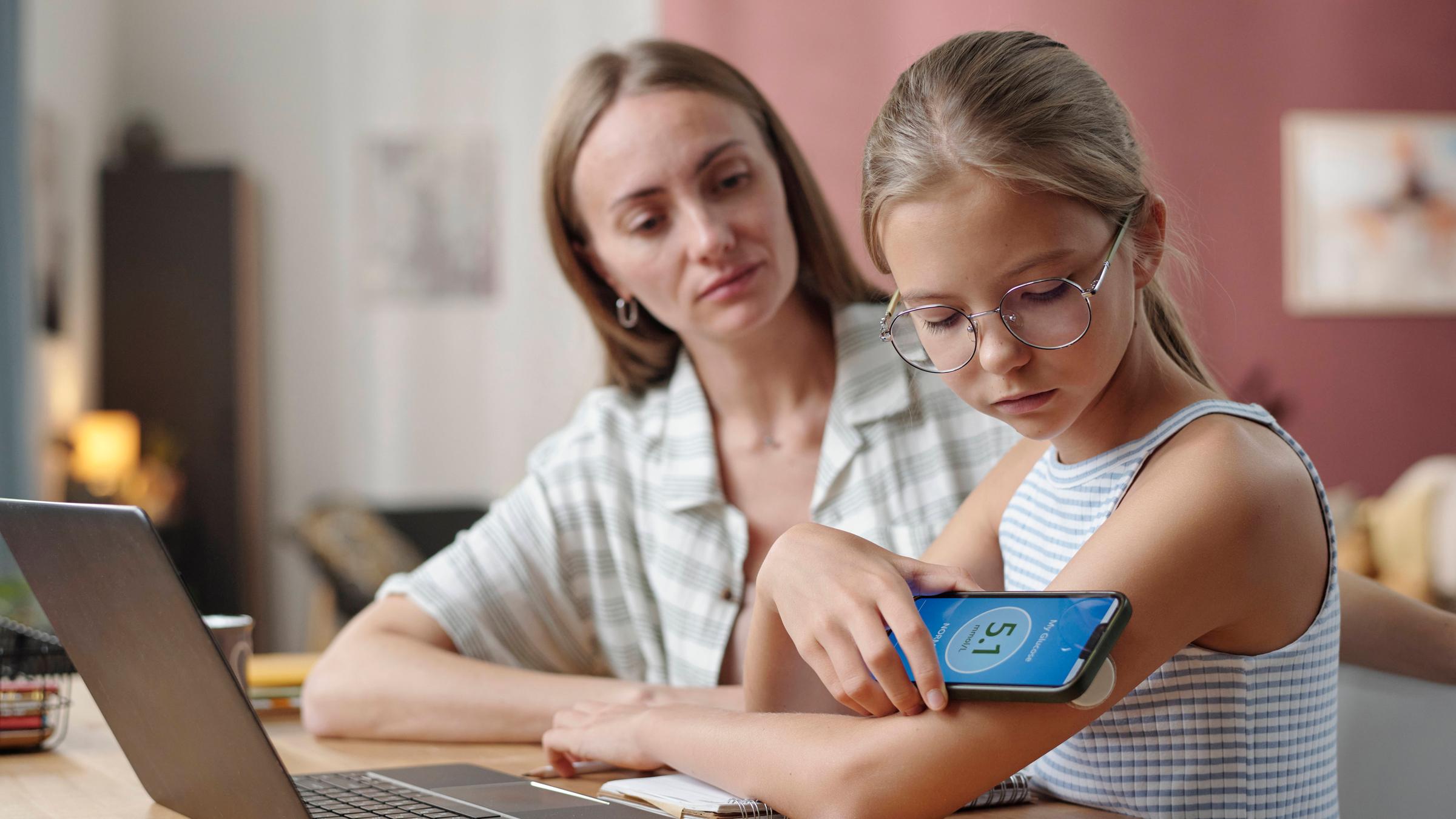 Eine Mutter sitzt mit ihrer Tochter am Tisch und unterstützt sie bei der Nutzung einer Diabetes-App. Auf dem Tisch steht ein Laptop.