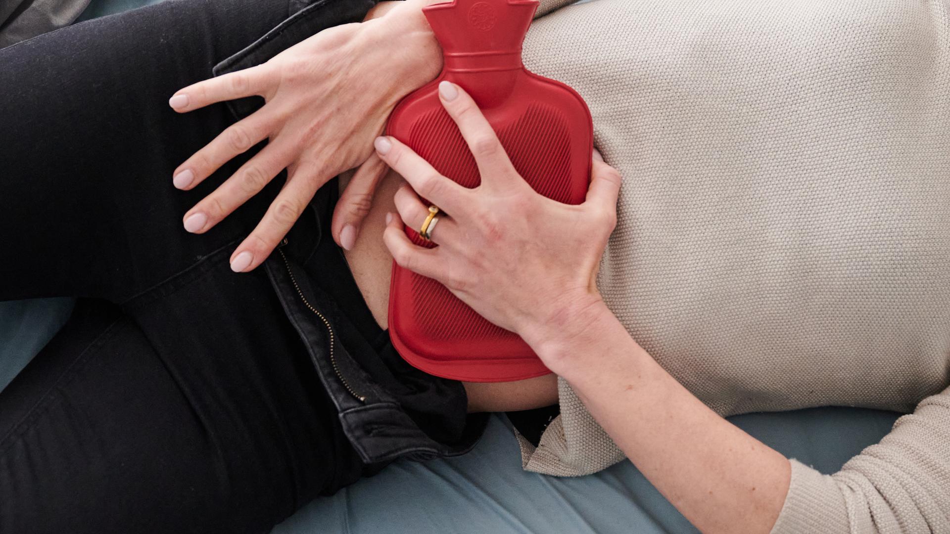 Eine Frau hält sich eine Wärmflasche an den Unterbauch, während sie im Bett liegt.
