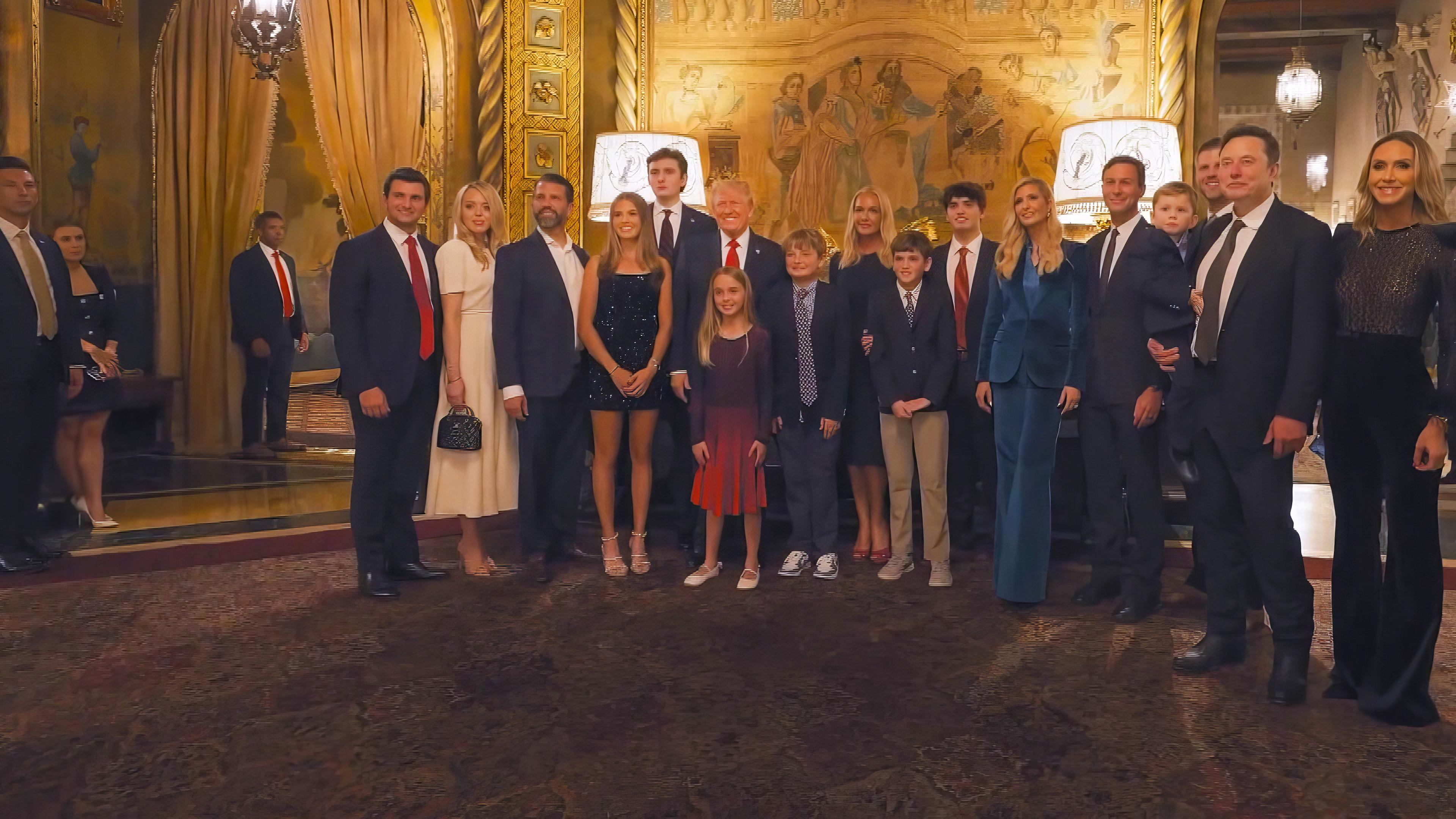 Donald Trump steht umringt von Familienmitgliedern und Elon Musk in einem prächtigen Foyer. Alle sind festlich gekleidet und posieren für ein Foto.