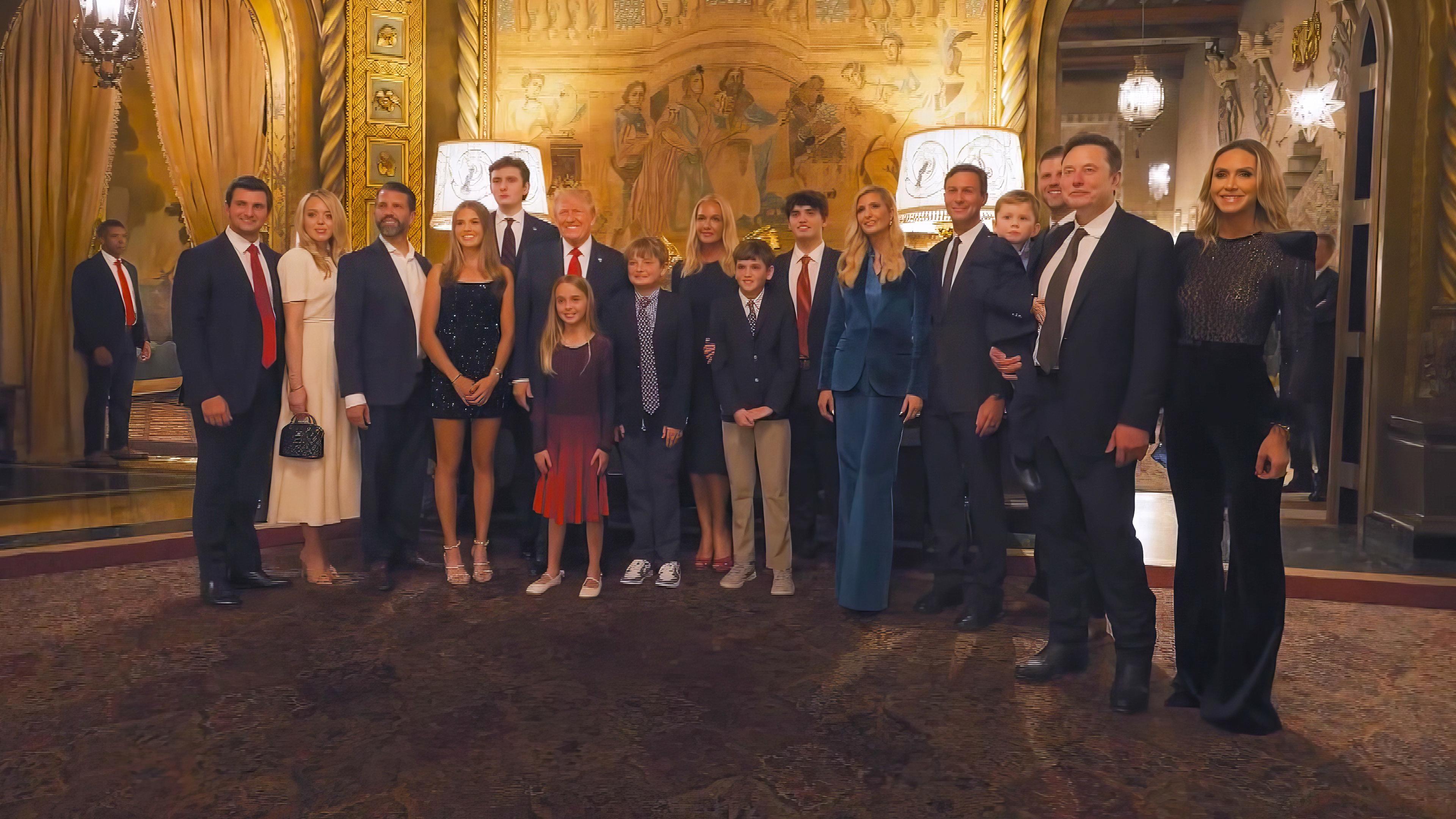 Donald Trump steht umringt von Familienmitgliedern und Elon Musk in einem prächtigen Foyer. Alle sind festlich gekleidet und posieren für ein Foto.