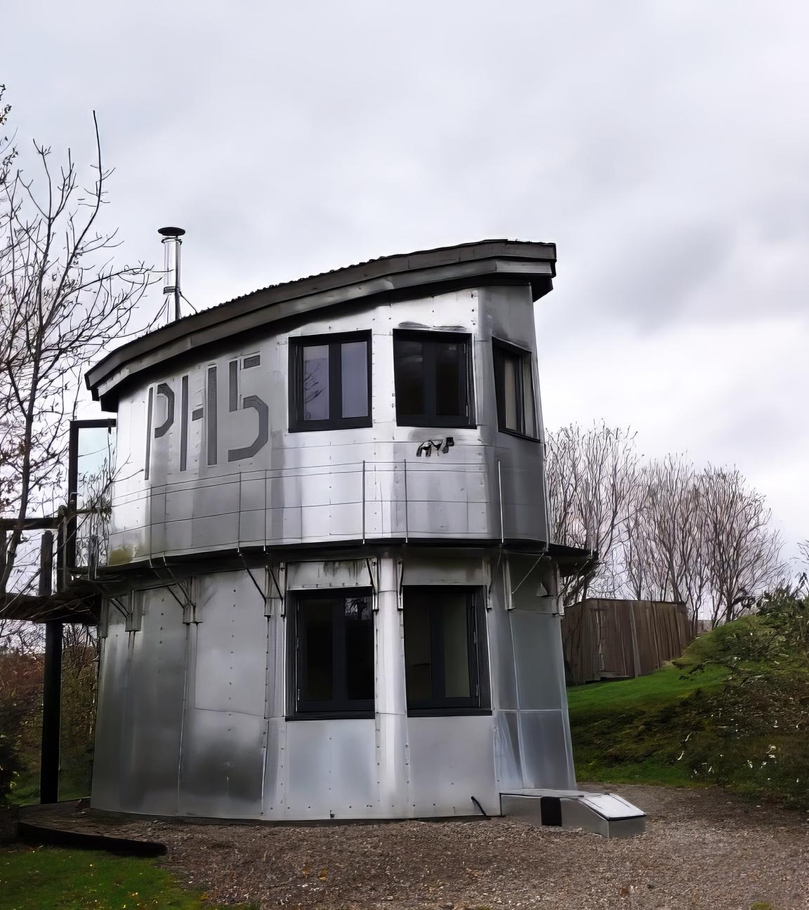 Ein silberner, zweistöckiger Wachturm mit großen Fenstern und der Nummer 'PH15' an der Seite steht an einem Weg im Grünen. Er ist umgeben von Bäumen und bewölktem Himmel